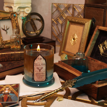 Load image into Gallery viewer, A bronze-colored candle with layers of white and teal wax, labeled 'Bellara', burning inside a large glass jar on a table with various items.