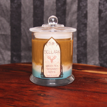 Load image into Gallery viewer, A bronze coloured candle labeled 'Bellara's Blend' with layers of white and teal wax, sitting on a wooden surface.