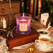 Load image into Gallery viewer, A pink and purple soy candle with the label reading "Deviant Dreamers - Lavender, Bergamot, White Tea, Cashmere - Planeshift Candles Custom Soy Candle" is placed on a wooden box amid flowers, tea leaves and books.