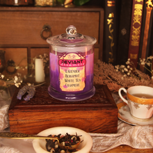 Load image into Gallery viewer, A pink and purple soy candle with the label reading "Deviant Dreamers - Lavender, Bergamot, White Tea, Cashmere - Planeshift Candles Custom Soy Candle" is placed on a wooden box amid flowers, tea leaves and books.