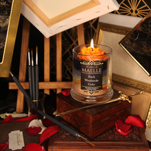 Load image into Gallery viewer, A lit orange wax rose sits on black, grey and orange tinted candle. The label reads "Planeshift Candles - Maelle - peach, woodsmoke - cedar - Clair Obscur expedition 33 inspired soy candle"
Hanging canvases and frames loom around the candle. Rose petals, paint brushes and a golden sword litter the floor.