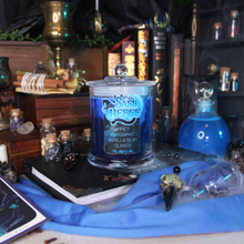Load image into Gallery viewer, A two tone blue soy candle with a label reading "SasiCheese - Honey - Woodsmoke - Vanilla Bean - Cloves - Planeshift Candles" sits on a black book surrounded by potion bottles, candles and gems.