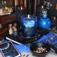 Load image into Gallery viewer, A two tone blue soy candle with a label reading "SasiCheese - Honey - Woodsmoke - Vanilla Bean - Cloves - Planeshift Candles" sits on a black book surrounded by potion bottles, candles and gems.