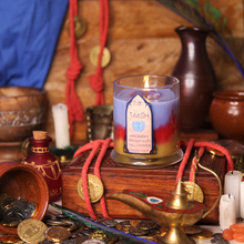 Load image into Gallery viewer, A candle with blue wax on top, red in the middle, and metallic gold at the bottom, labeled 'Taash's Blend'. It is set in a clear glass jar and placed on a wooden surface with decorative items around it.