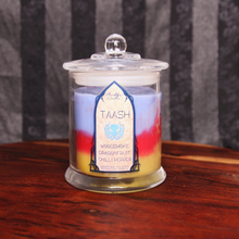 Load image into Gallery viewer, Our Dragon Age Taash inspired candle in a clear glass jar with lid and ight blue, red and golden wax. Label reads "Taash - Woodsmoke, Dragonfruit, Chillipepper"