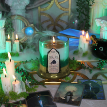 Load image into Gallery viewer, A clear glass jar with a burning  white, green and golden candle inside, surrounded by smoke, various other candles and tarot cards on the table.
