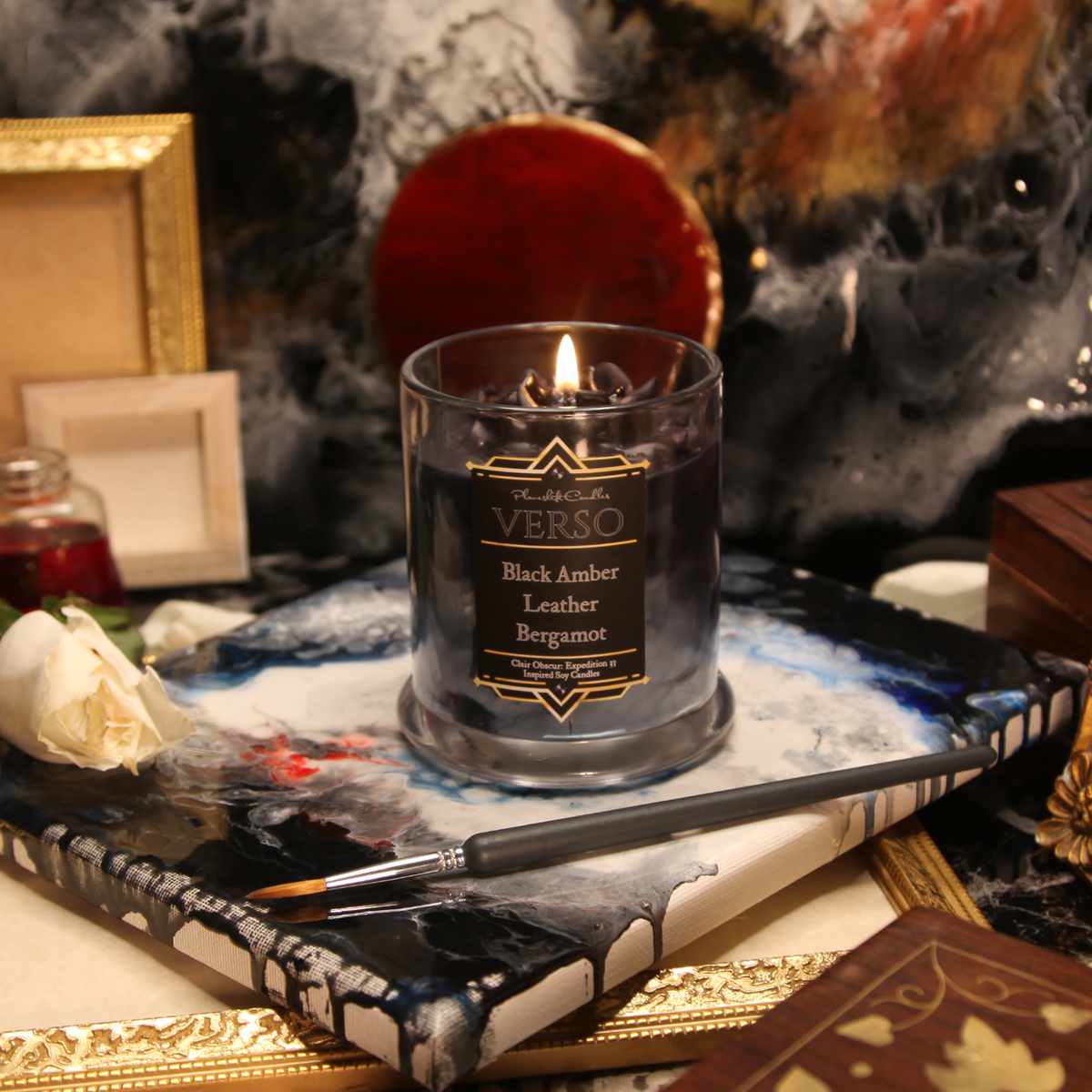 A soy candle with a purple rose on a black, grey and violet candle. The label reads "Planeshift Candles - Verso - Black Amber, Leather, Bergamot - Clair Obscur: Expedition 33 inspired Soy Candle" 